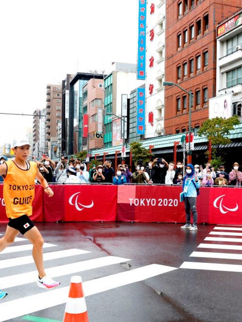 A male runner and his guide compete in the marathon at Tokyo 2020. People are taking photos as they compete.
